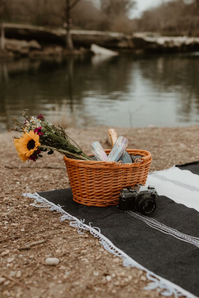 Picnic basket with a bouquet of flowers, two champagne glasses, and a vintage film camera arranged on a blanket.