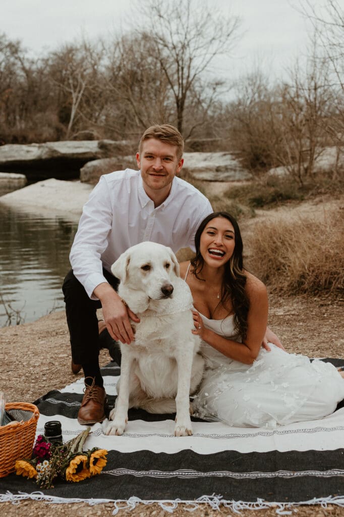 A couple sitting on a picnic blanket with their dog, smiling, at McKinney Falls State Park.