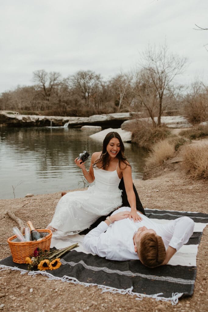 A couple sitting on a picnic blanket, smiling and holding a vintage film camera at McKinney Falls State Park.