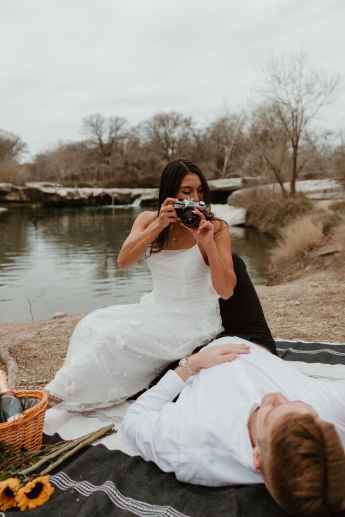 A couple sitting on a picnic blanket, smiling and holding a vintage film camera at McKinney Falls State Park.