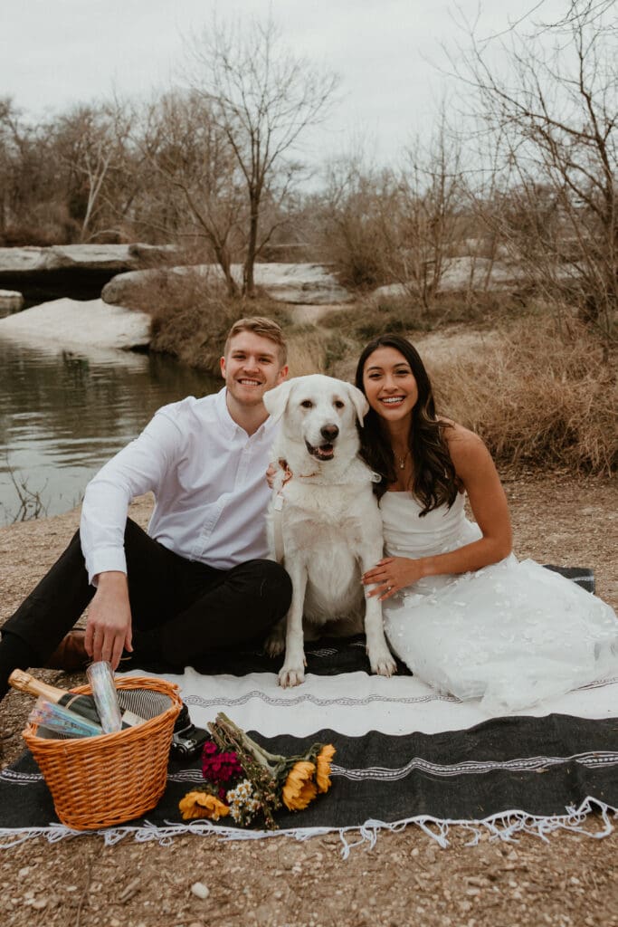 A couple sitting on a picnic blanket with their dog during an engagement photoshoot at McKinney Falls State Park.