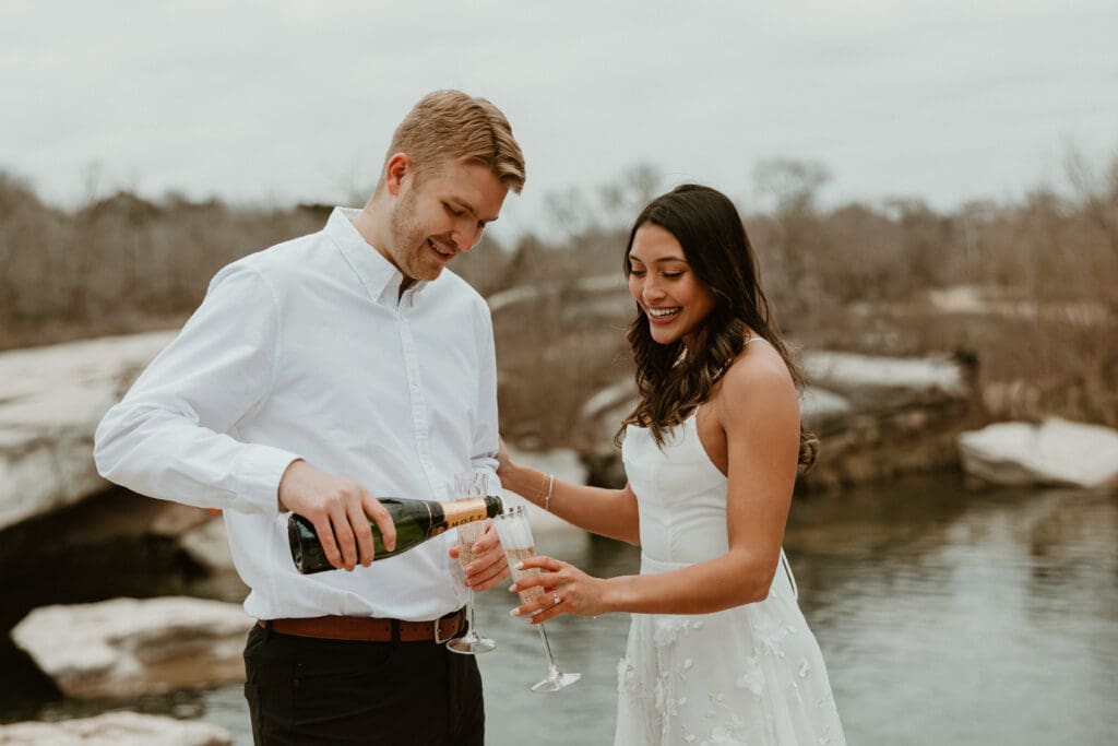 An engaged couple celebrating with a toast overlooking the cliffs at McKinney Falls State Park in Austin Texas.