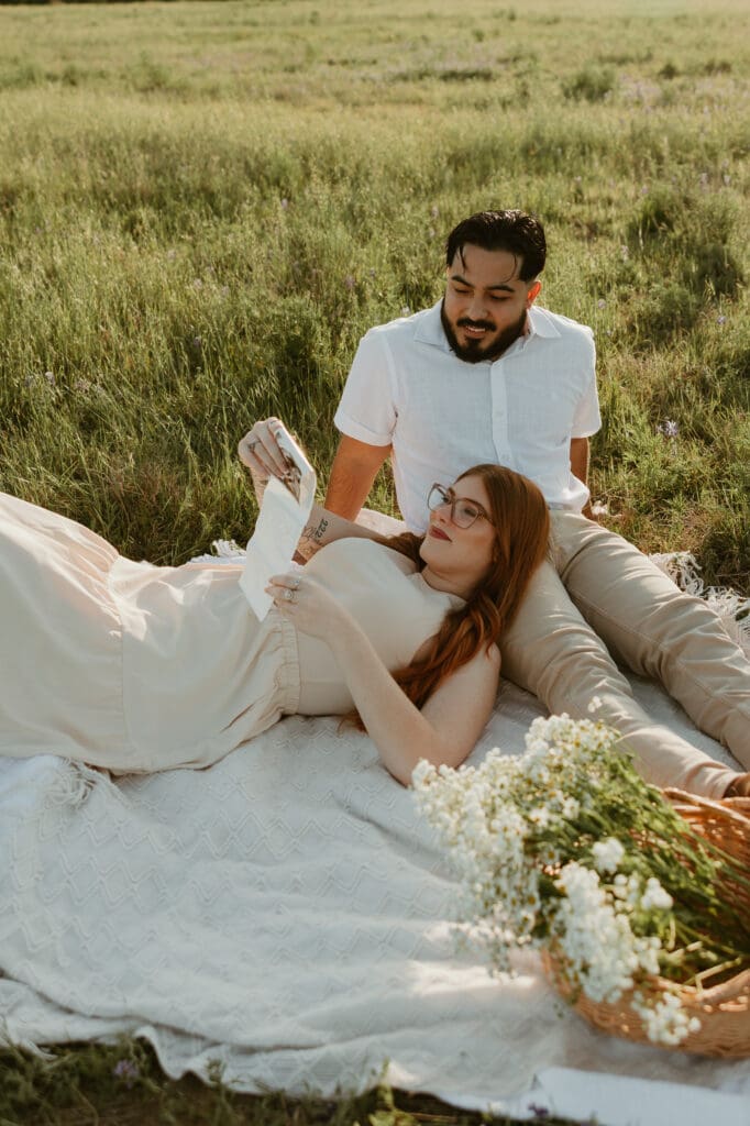 Couple sitting together on a picnic blanket, showcasing baby announcement props with dreamy sunset lighting.

