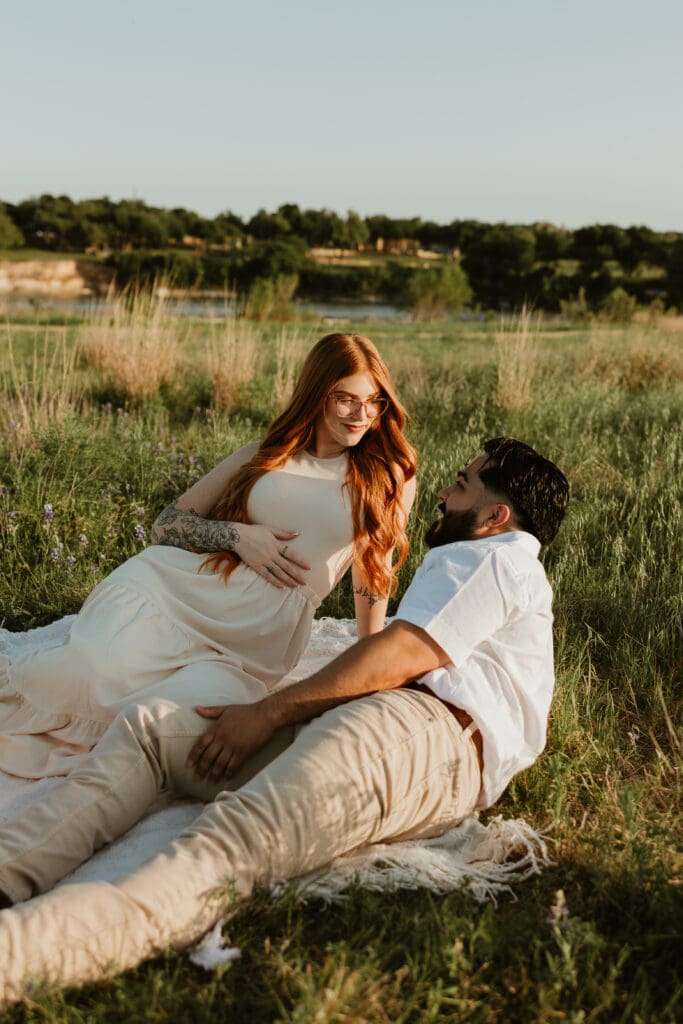 Couple sitting together on a picnic blanket, holding baby bump during photo session in Cedar Park, Texas.
