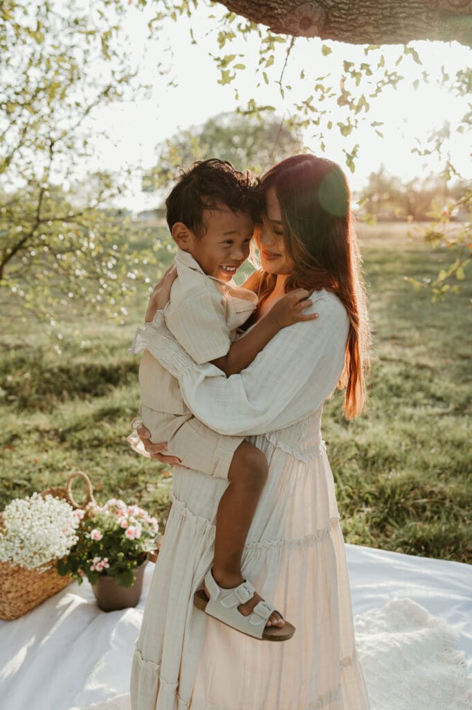 Mother cuddling her toddler son close, both wrapped in a warm golden light with soft trees in the background.