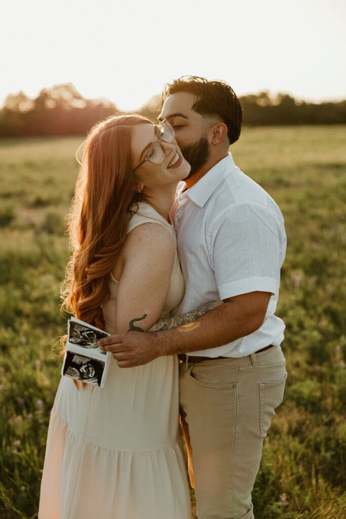 Pregnant couple standing in a field of wildflowers with an ultrasound image with a golden light silhouette filling the background. 