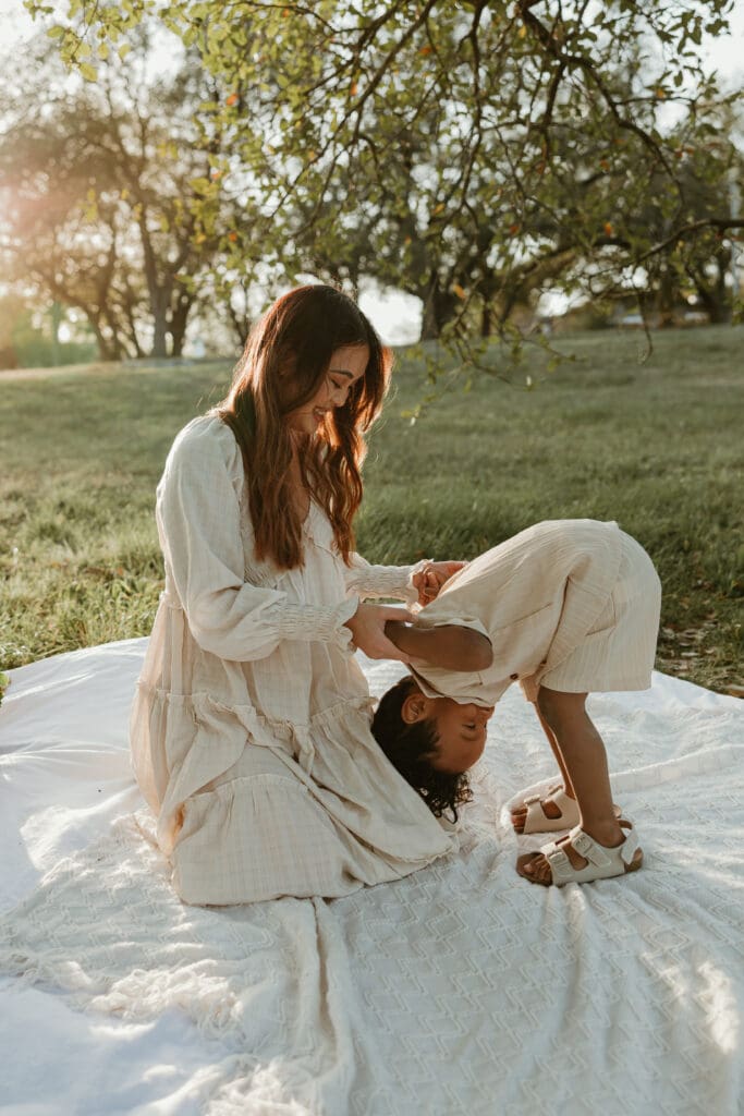 Toddler laughing joyfully as his mom tickles him, both framed by glowing sunlight filtering through the trees.