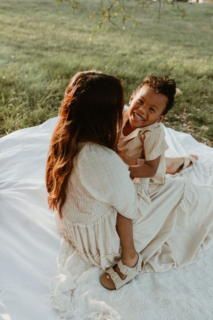 Toddler laughing joyfully as his mom tickles him, both framed by glowing sunlight filtering through the trees.