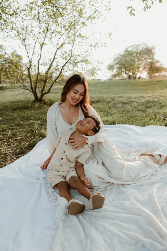 Mother cuddling her toddler son close, both wrapped in a warm golden light with soft trees in the background.