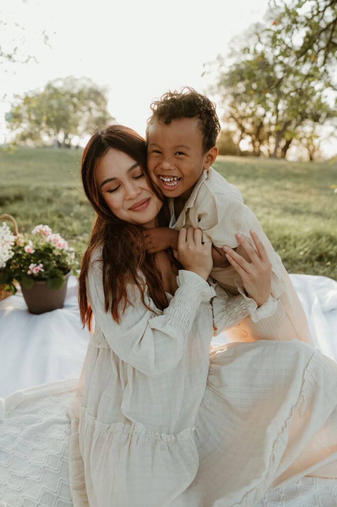 Three-year-old boy giggling while his mom crouches beside him, both bathed in soft golden light at a scenic park in North Austin.