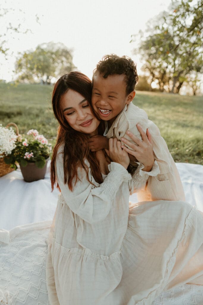 Mother cuddling her toddler son close, both wrapped in a warm golden light with soft trees in the background.