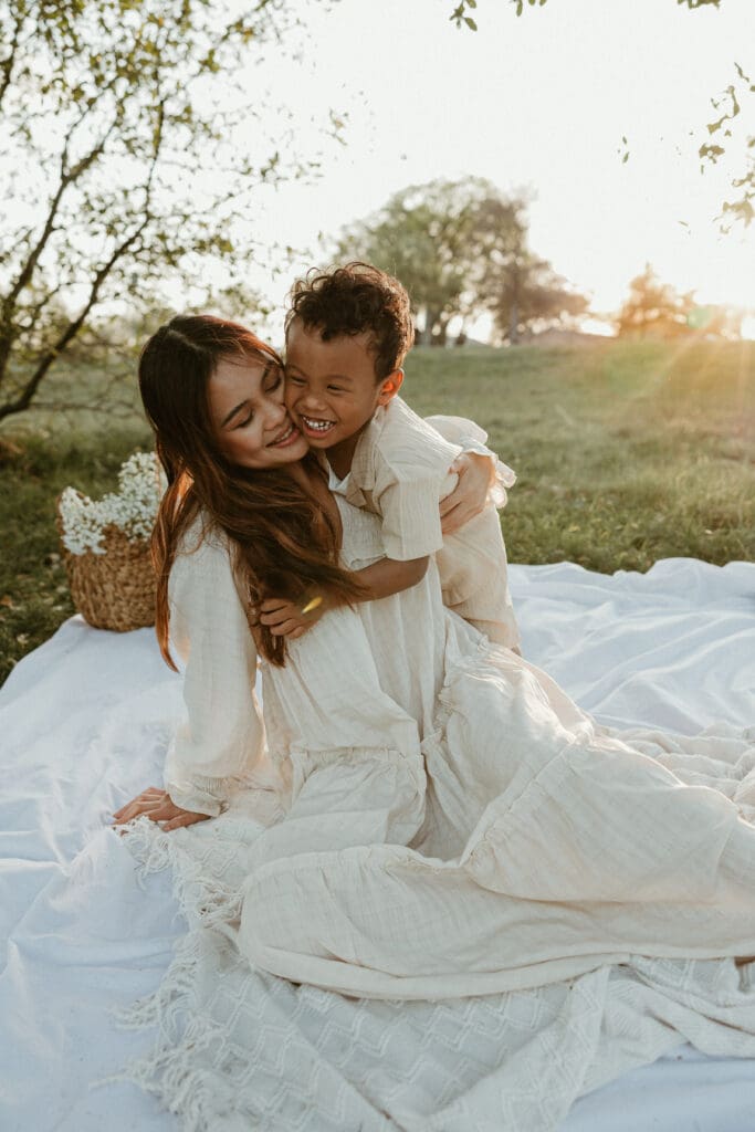 Three-year-old boy giggling while his mom crouches beside him, both bathed in soft golden light at a scenic park in North Austin.