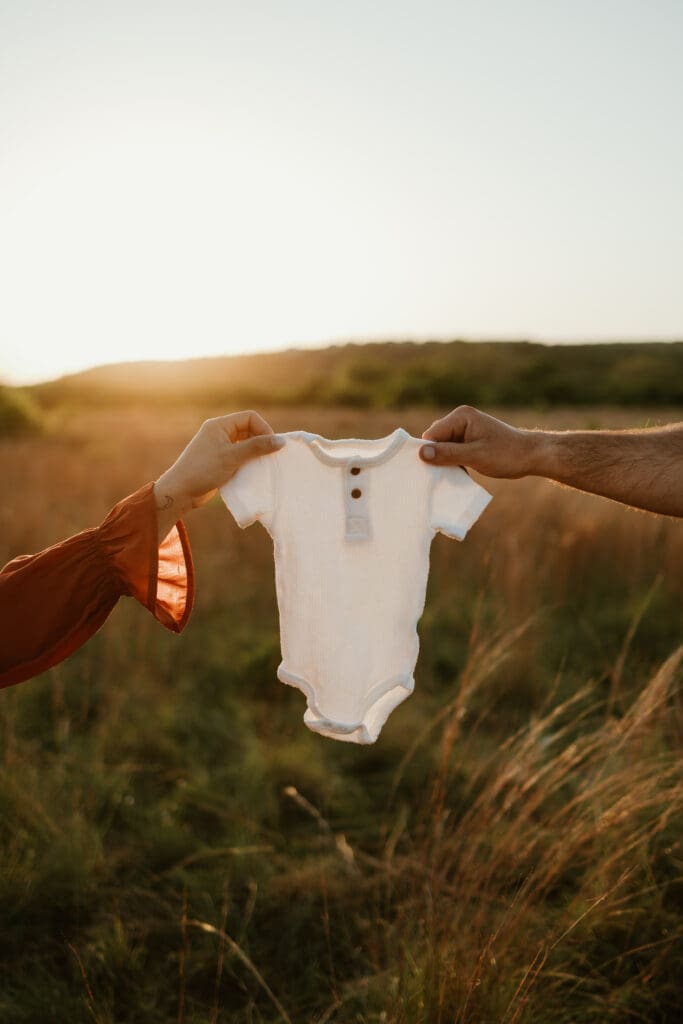 Expecting parents hold newborn onesie in open field during golden hour at Common Fords Ranch in Austin. 