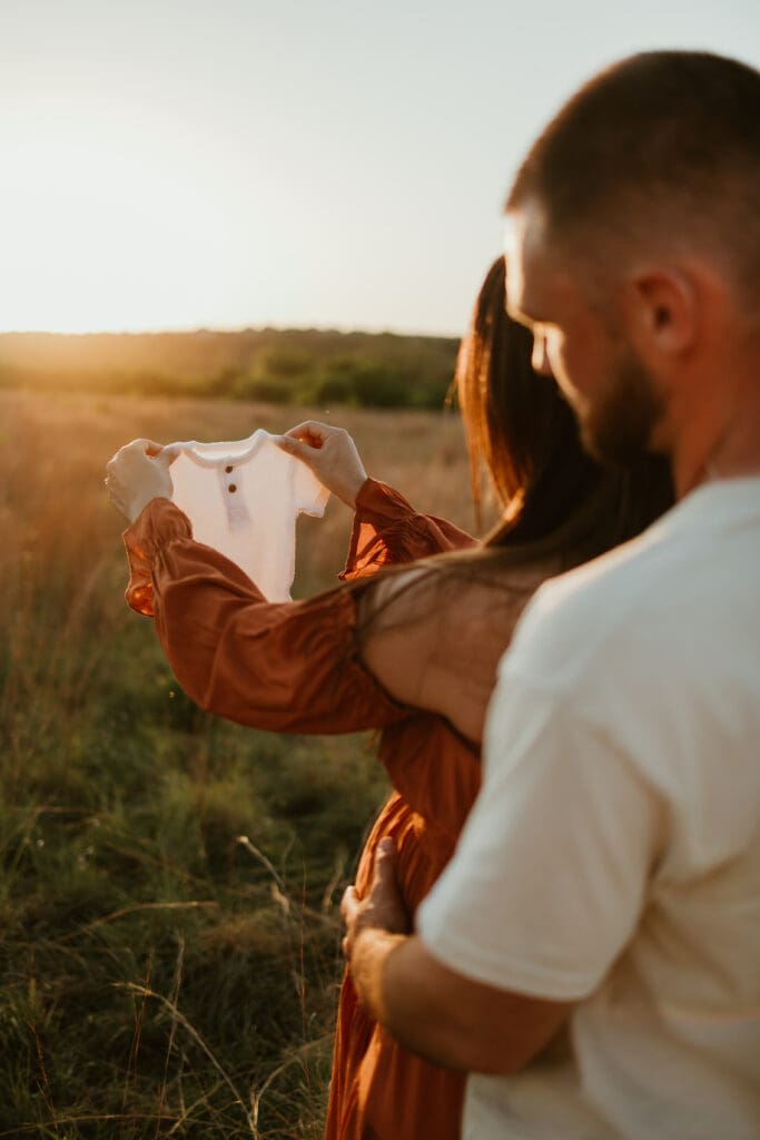Expecting parents hold newborn onesie during golden hour photoshoot at Common Fords Ranch in Austin. 