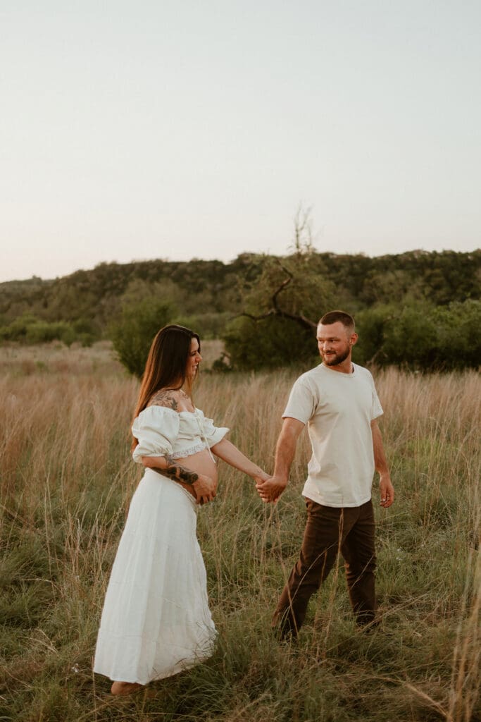 Expecting parents hold hands in open field during golden hour at Common Fords Ranch in Austin. 